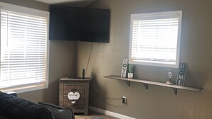 Un salon doté d’un téléviseur à écran plat fixé au mur, d’un meuble en bois sur lequel est posé un panneau indiquant « Bienvenue », et de deux fenêtres équipées de stores.