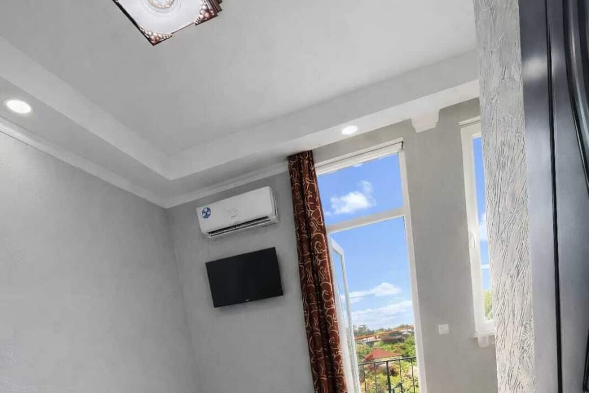 A modern bedroom with a bed, a small bedside table, a TV mounted on the wall, an air conditioning unit, and a window with curtains.