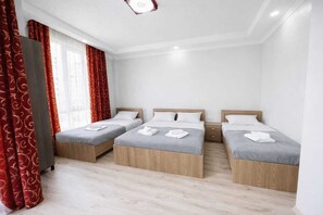 Ein Hotelzimmer mit drei Einzelbetten, einem großen Fenster mit roten Vorhängen und Holzboden.