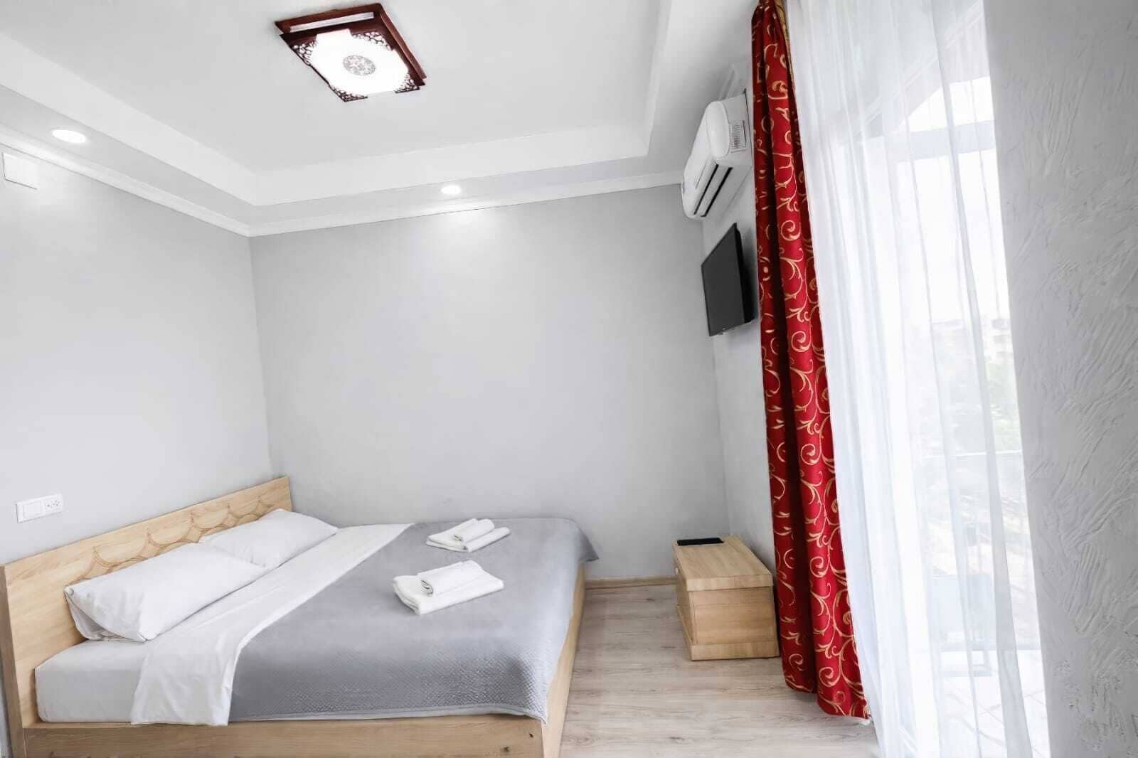 Un dormitorio con cama, mesita de noche, televisor montado en la pared y ventana con cortinas rojas.