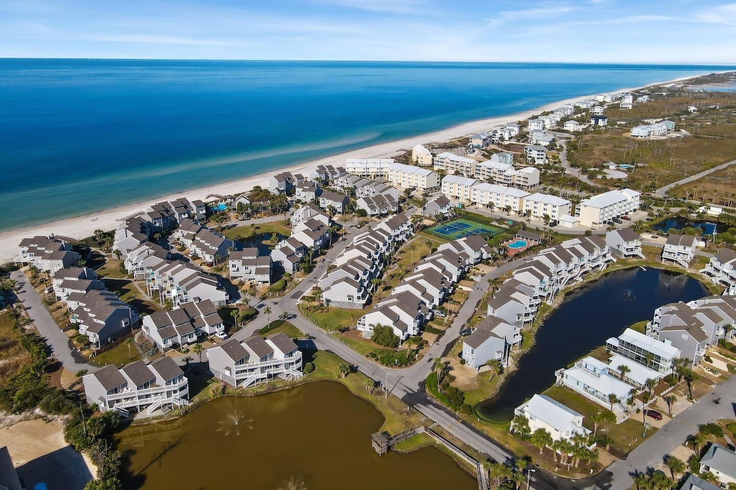 Vue aérienne d’une zone résidentielle côtière, avec des maisons à plusieurs étages, une plage et un petit lac.