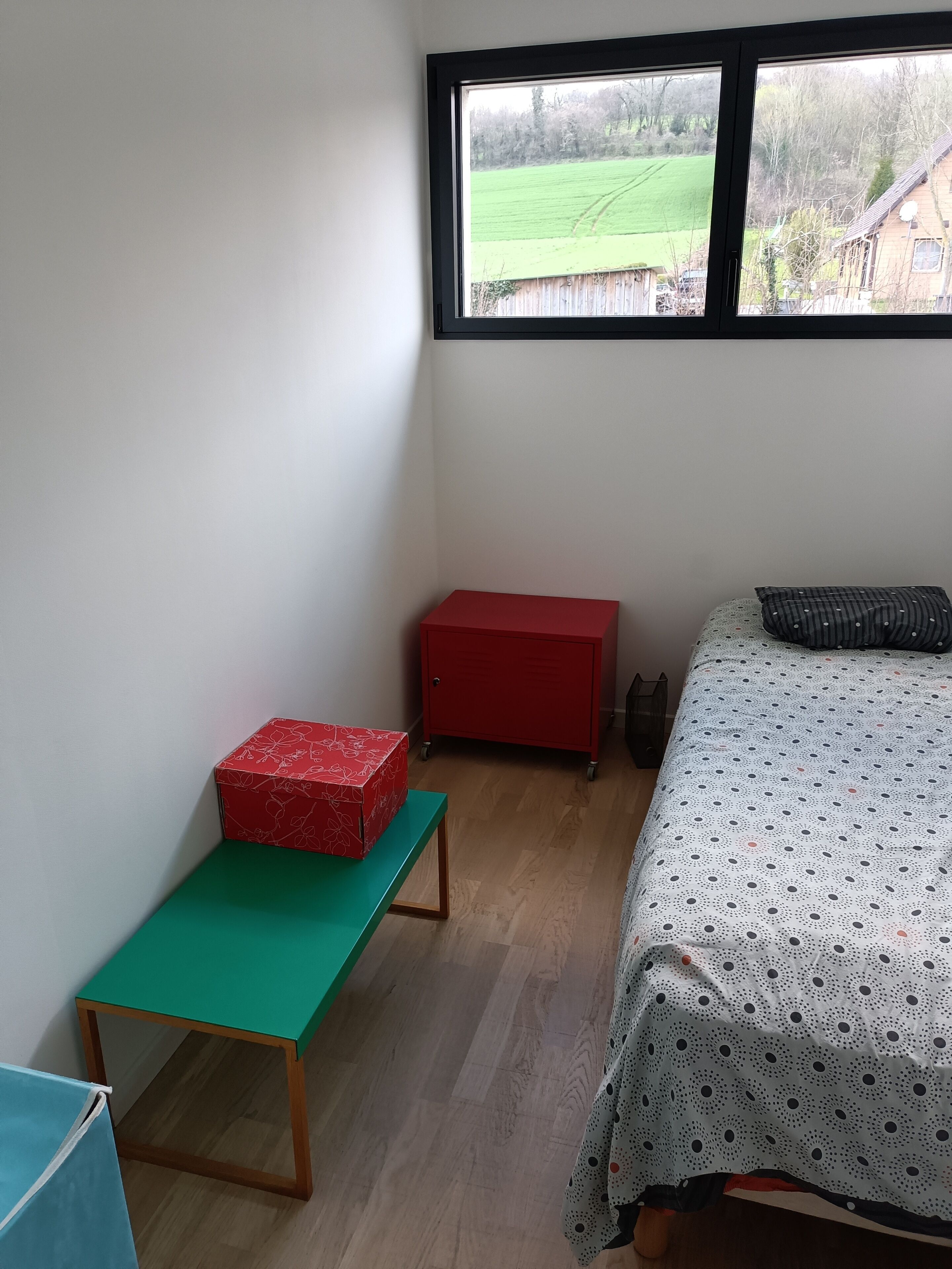 A small, sparsely furnished room with a single bed, a green table, a red storage unit, and a window overlooking a green field.