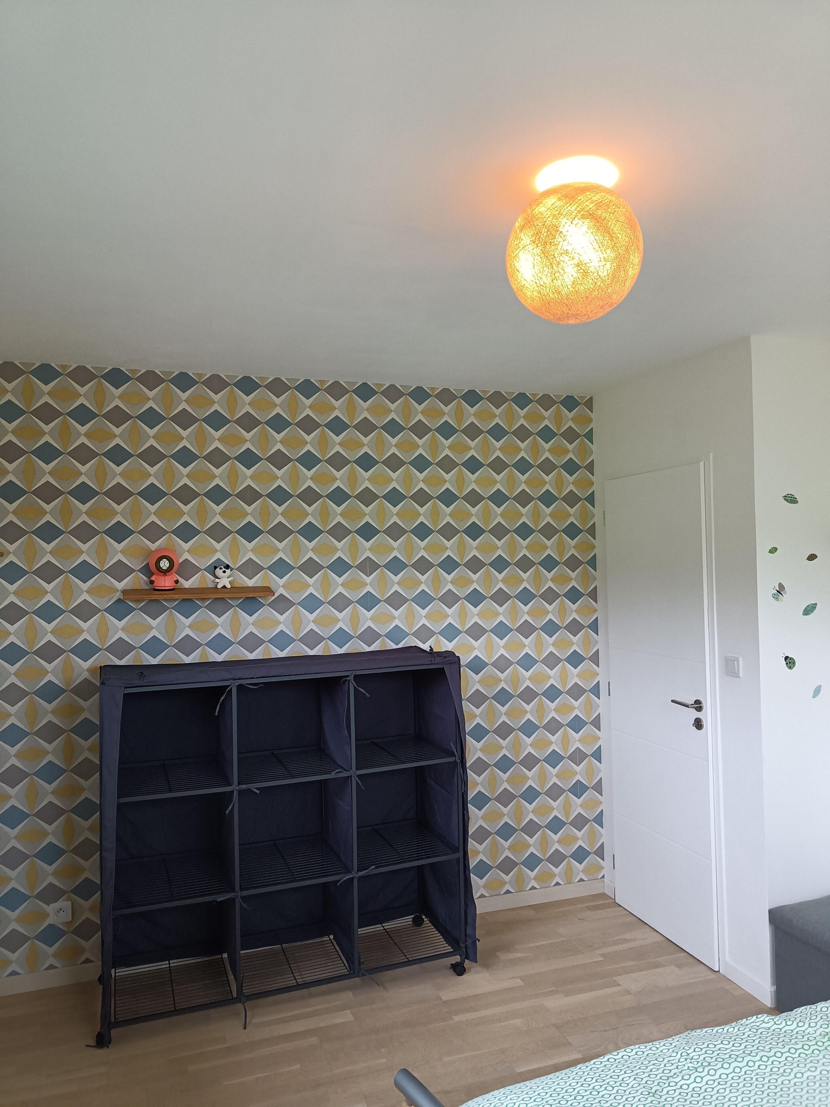 A room with a patterned wallpaper, a black shelf unit, a door, and a hanging light fixture.
