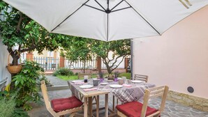Ein Essbereich im Freien mit einem Tisch für vier Personen, umgeben von Holzstühlen mit roten Kissen, unter einem großen weißen Sonnenschirm.