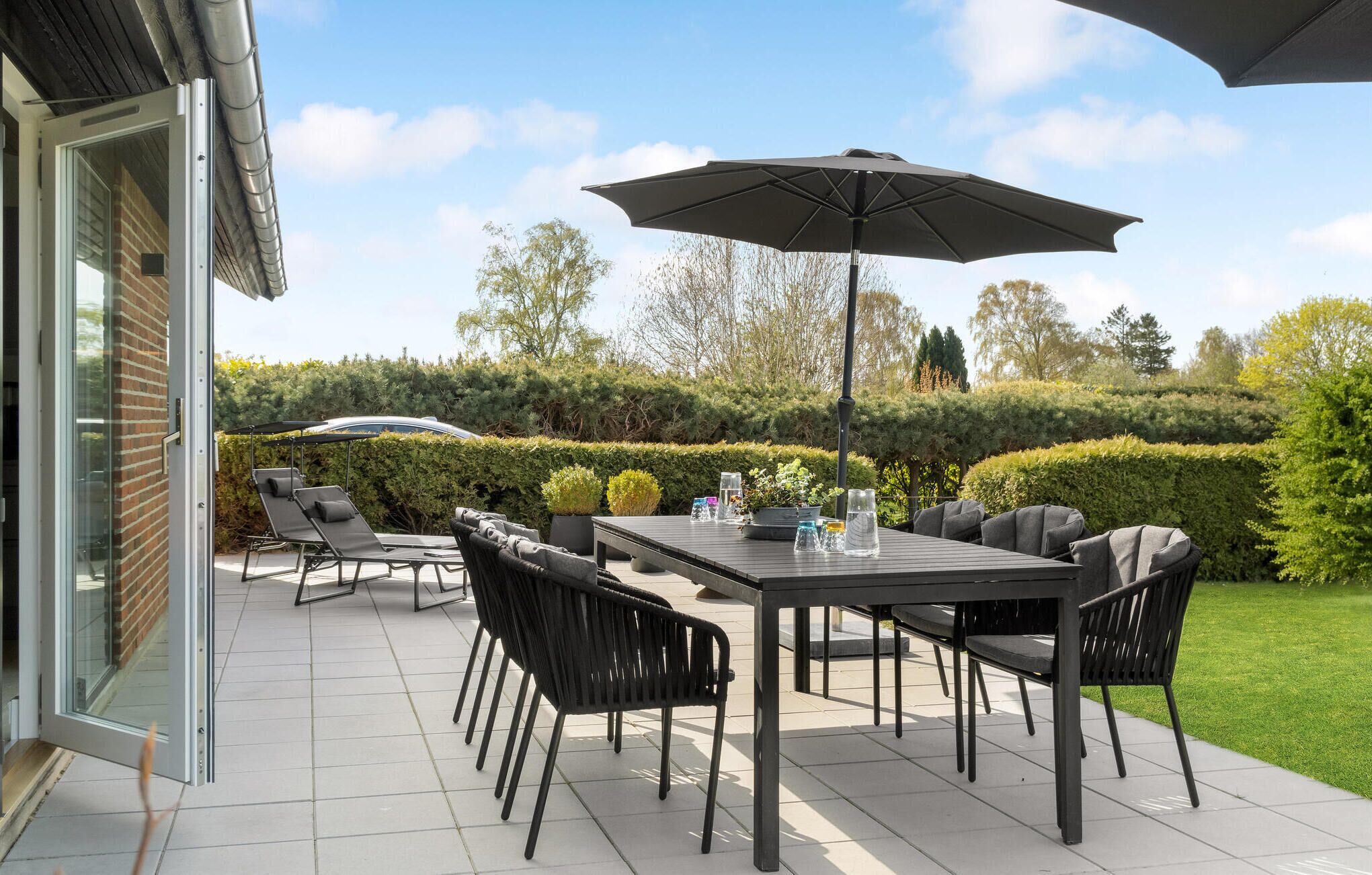 A patio with a table and chairs, a large umbrella, and a view of a hedge and trees.