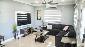 Una sala de estar moderna con un sofá seccional, un televisor de pantalla plana, un ventilador de techo y un amplio ventanal con persianas.