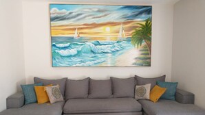 A grey sectional sofa with patterned cushions, a glass coffee table, and a painting of a sunset over the ocean with sailboats.