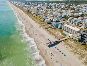 An aerial view of a beachside town with a long sandy beach, a pier, and a row of buildings including a hotel with a pool.