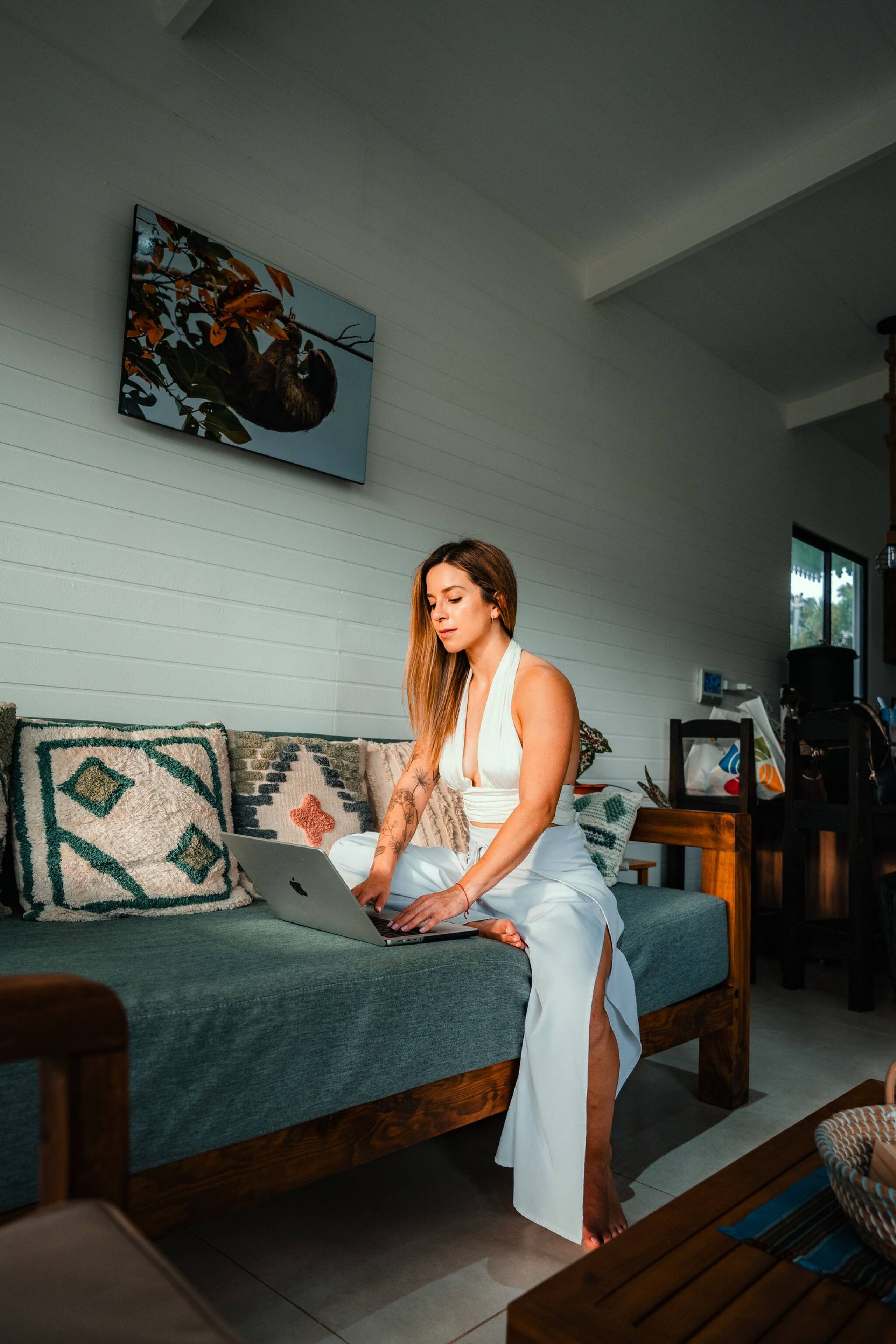 A person sitting on a sofa with a laptop, in a room with a wooden floor, a painting on the wall, and a window.
