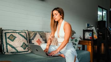 A person sitting on a sofa with a laptop, in a room with a wooden floor, a painting on the wall, and a window.