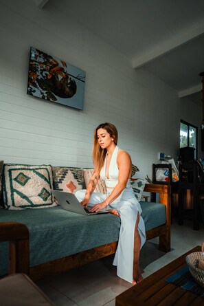 A person sitting on a sofa with a laptop, in a room with a wooden floor, a painting on the wall, and a window.
