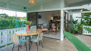 Un espace extérieur abrité, aménagé pour les repas, avec une table ronde en bois et des chaises en osier, un luminaire suspendu, et une vue sur la cuisine et l’espace de vie adjacents.