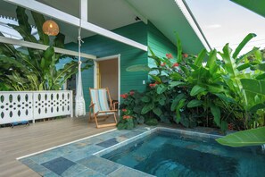 A pool area with a wooden deck, a lounge chair, and lush tropical plants.
