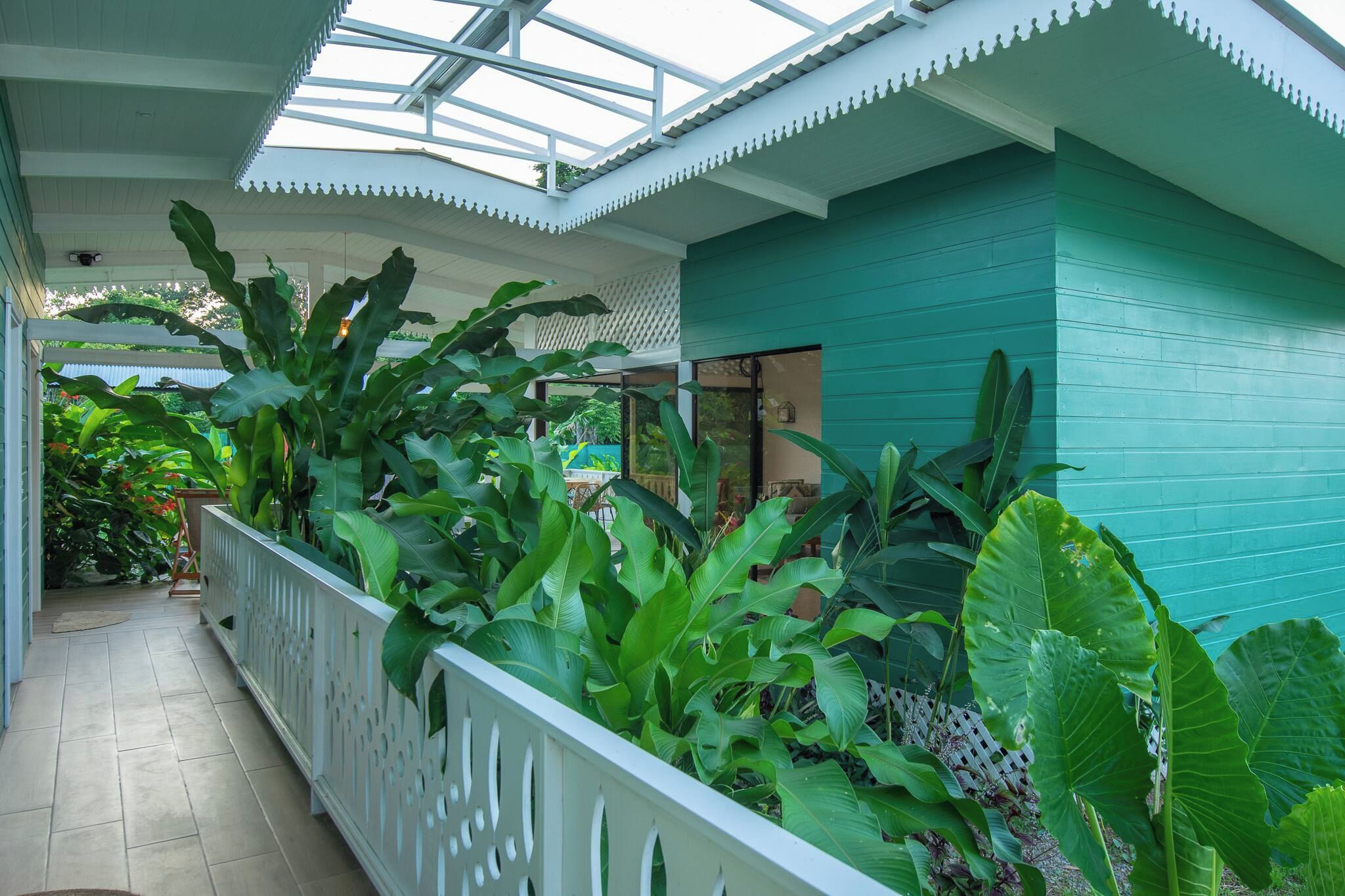 A covered walkway with a white railing, surrounded by lush green plants and a teal-colored wall.