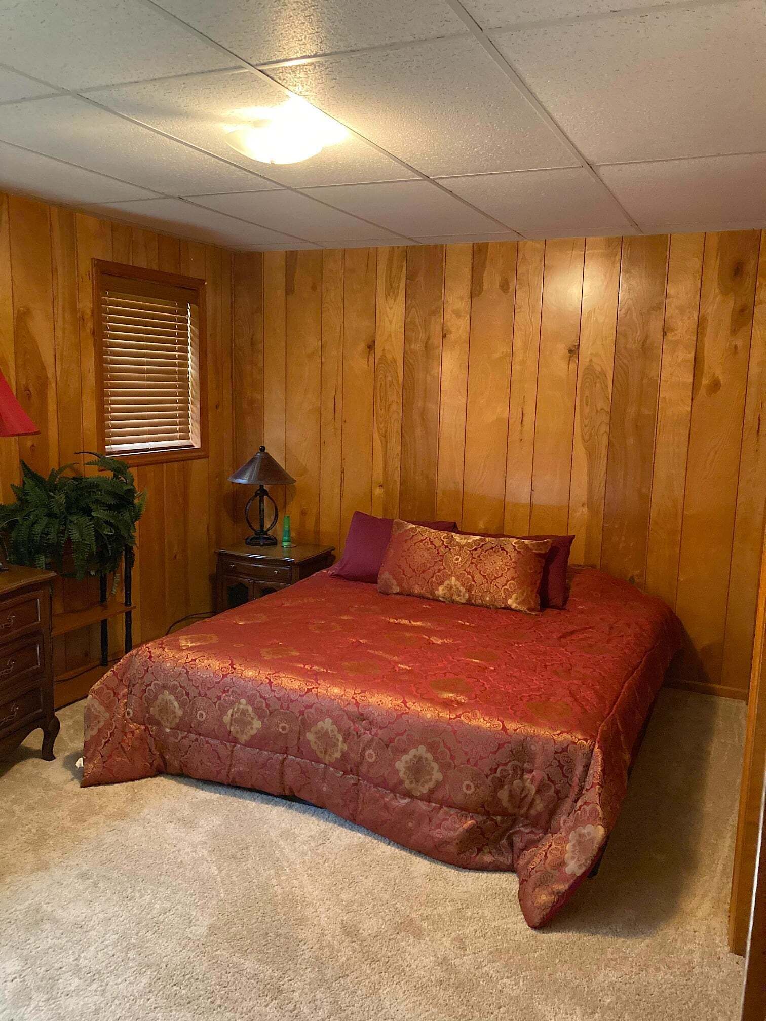 A bedroom with a wooden paneled wall, a bed with a red bedspread, a nightstand with a lamp, and a window with blinds.