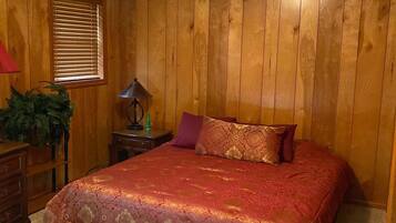 A bedroom with a wooden paneled wall, a bed with a red bedspread, a nightstand with a lamp, and a window with blinds.