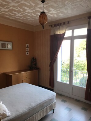 Une chambre à coucher comprenant un lit, une commode, un luminaire au plafond, une fenêtre avec des rideaux et une vue sur la verdure à l’extérieur.