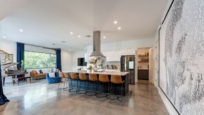 A spacious living area with a piano, a blue sofa, and a kitchen with a large island. - Modern Austin home w private pool and spa, Minutes to Lake Austin & Lake Travis! (Austin)