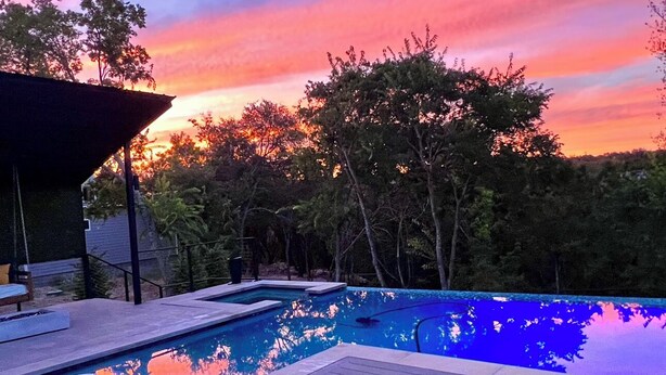 A swimming pool with lounge chairs and a deck, surrounded by trees and a sunset sky. - Modern Austin home w private pool and spa, Minutes to Lake Austin & Lake Travis! (Austin)