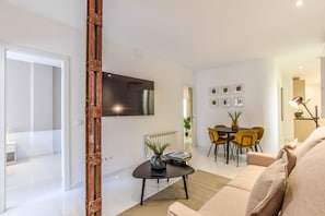 A modern living room with a flat-screen TV mounted on the wall, a coffee table with a vase and remote control, a sofa, and a dining area with chairs and a table.