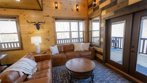 A cozy room with a brown leather sofa, a round coffee table, and a wooden wall with a deer head mounted above a window.