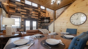 A dining area with a wooden table set for four, a clock on the wall, and a staircase leading to an upper level.