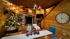 A cozy cabin interior with a wooden interior, a fireplace, a Christmas tree, a clock, and a toy train on the table.