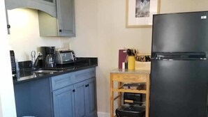A compact kitchen with grey cabinets, a black refrigerator, a coffee maker, and a trash bin. - Sheepscot Harbour Studio #215 (Edgecomb)