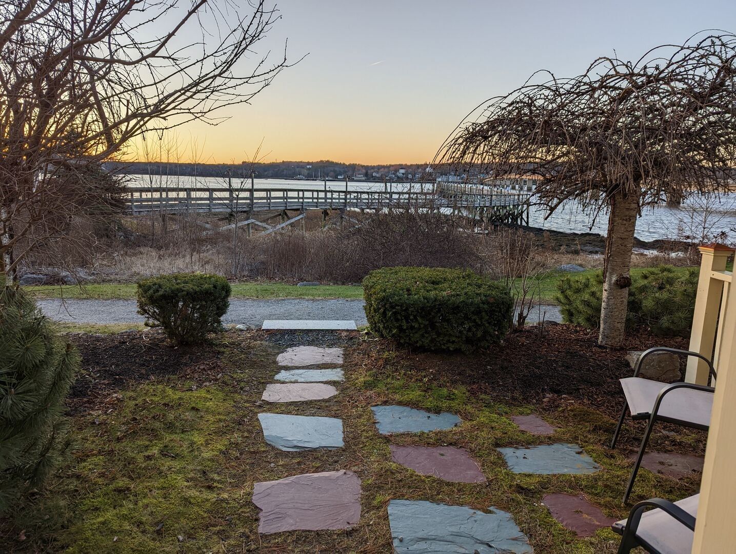 Un sentier de pierres mène à une terrasse en bois qui surplombe une rivière, avec un pont en arrière-plan.