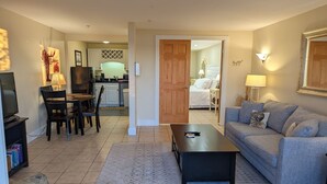 A living room with a sofa, a coffee table, and a TV. There is a dining area with chairs and a table. A kitchen is visible in the background. - Waterfront king studio with kitchen Sheepscot Harbour Studio #205 (Edgecomb)