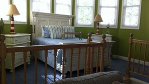 A bedroom with a bed, bedside tables, a nightstand, a wooden crib, and a window with blinds. - Sheepscot Harbour Studio #308 (Edgecomb)