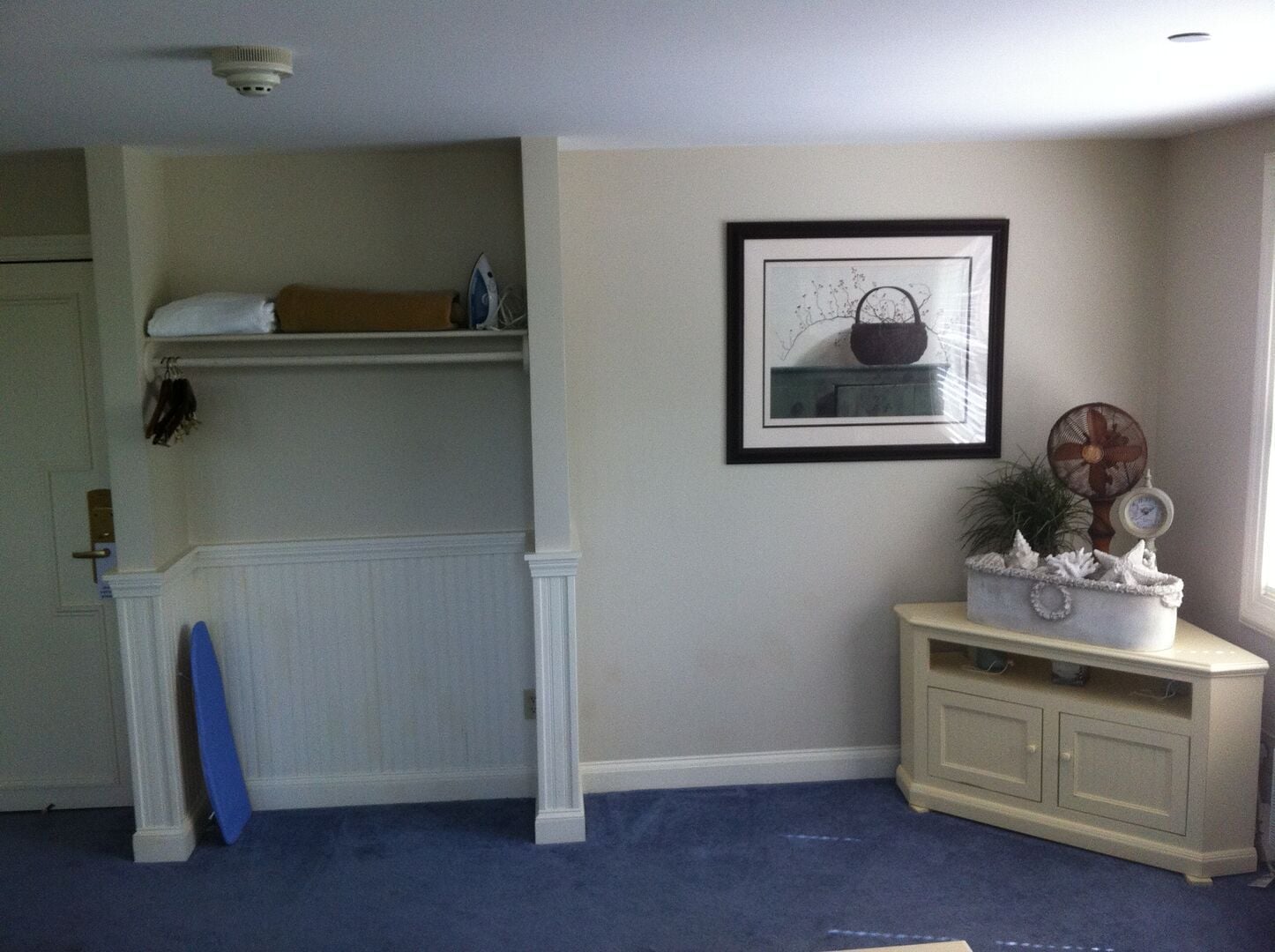 A room with a white door, a blue surfboard leaning against the wall, a framed picture above a white cabinet, and a ceiling fan on the right.