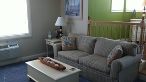 A living room with a striped sofa, a side table with a lamp, a framed picture on the wall, and a window with blinds. - Sheepscot Harbour Studio #308 (Edgecomb)