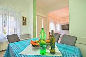 Een eettafel met een blauwe tafelkleed, twee stoelen, een waterfles, een fles wijn en een schaal fruit.