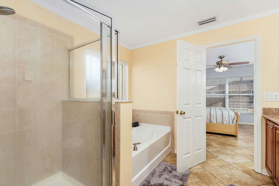 Ein Badezimmer mit Duschkabine aus Glas, einer Badewanne und einer Tür zu einem Schlafzimmer mit Bett und Deckenventilator.