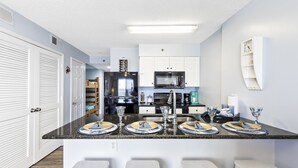 Een moderne keuken met witte kasten, een zwarte koelkast en een magnetron. Er staan drie barkrukken bij de aanrecht en een spoelbak met een kraan.