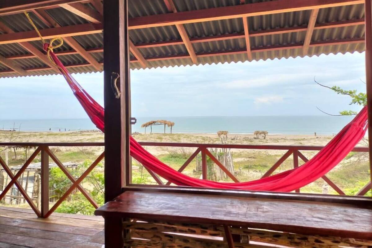Una terraza de madera con una hamaca roja, una mesa de madera y vistas a la playa y al océano.