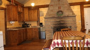 Una cocina acogedora de estilo cabaña, con armarios de madera, chimenea de piedra y una mesa de comedor cubierta con mantel a cuadros.