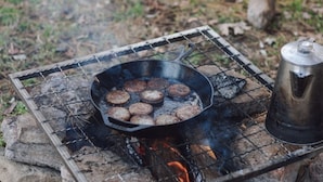 A portable grill with a cast iron skillet containing burgers, a metal pot, and a wire mesh grill over a fire. - Two cozy pods with water tower (Kingston)