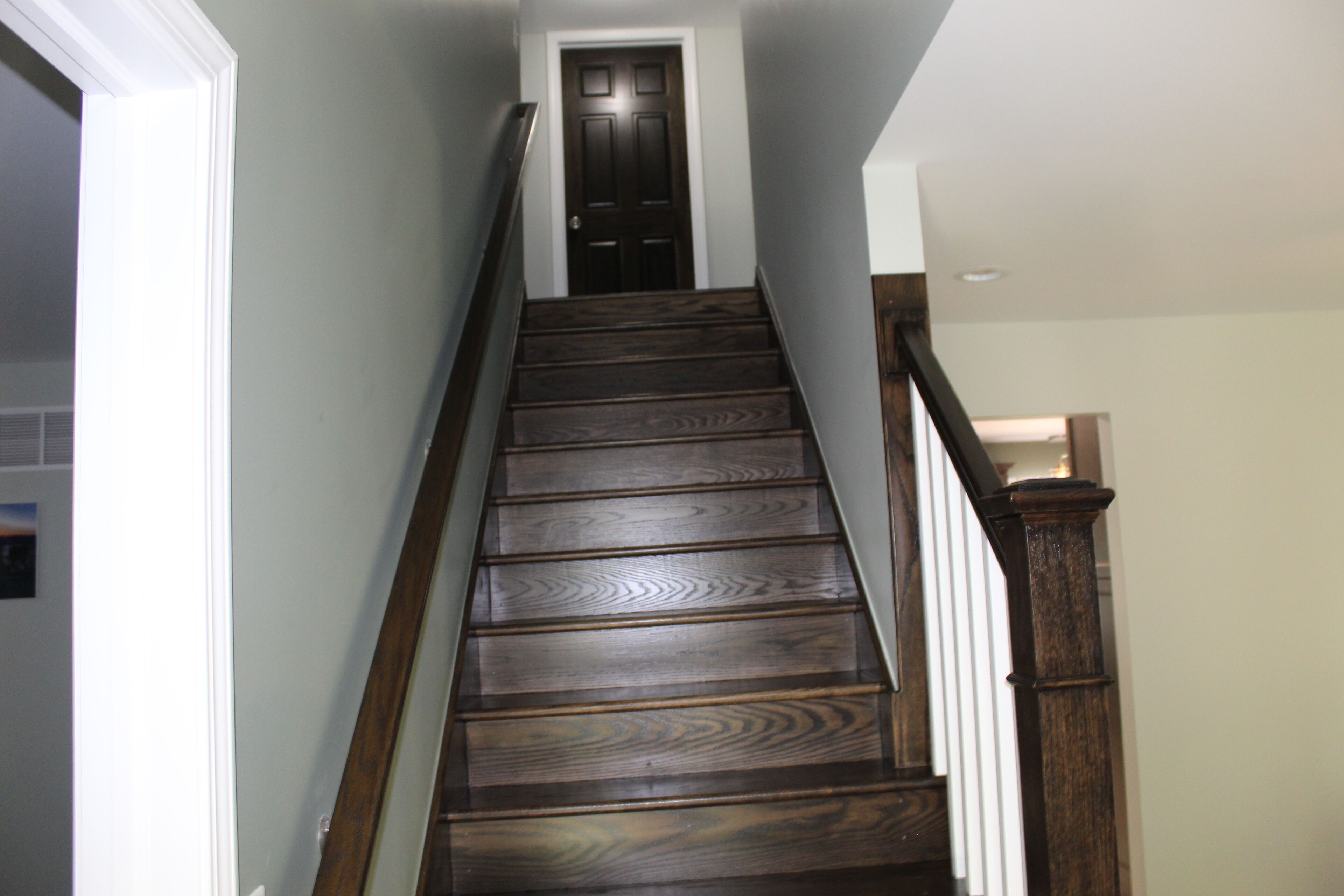 Un escalier en bois à la teinte sombre, avec des rampes blanches et une porte fermée en haut.