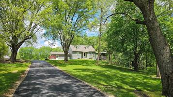 A two-story house with a front porch, surrounded by lush green trees and a paved driveway.