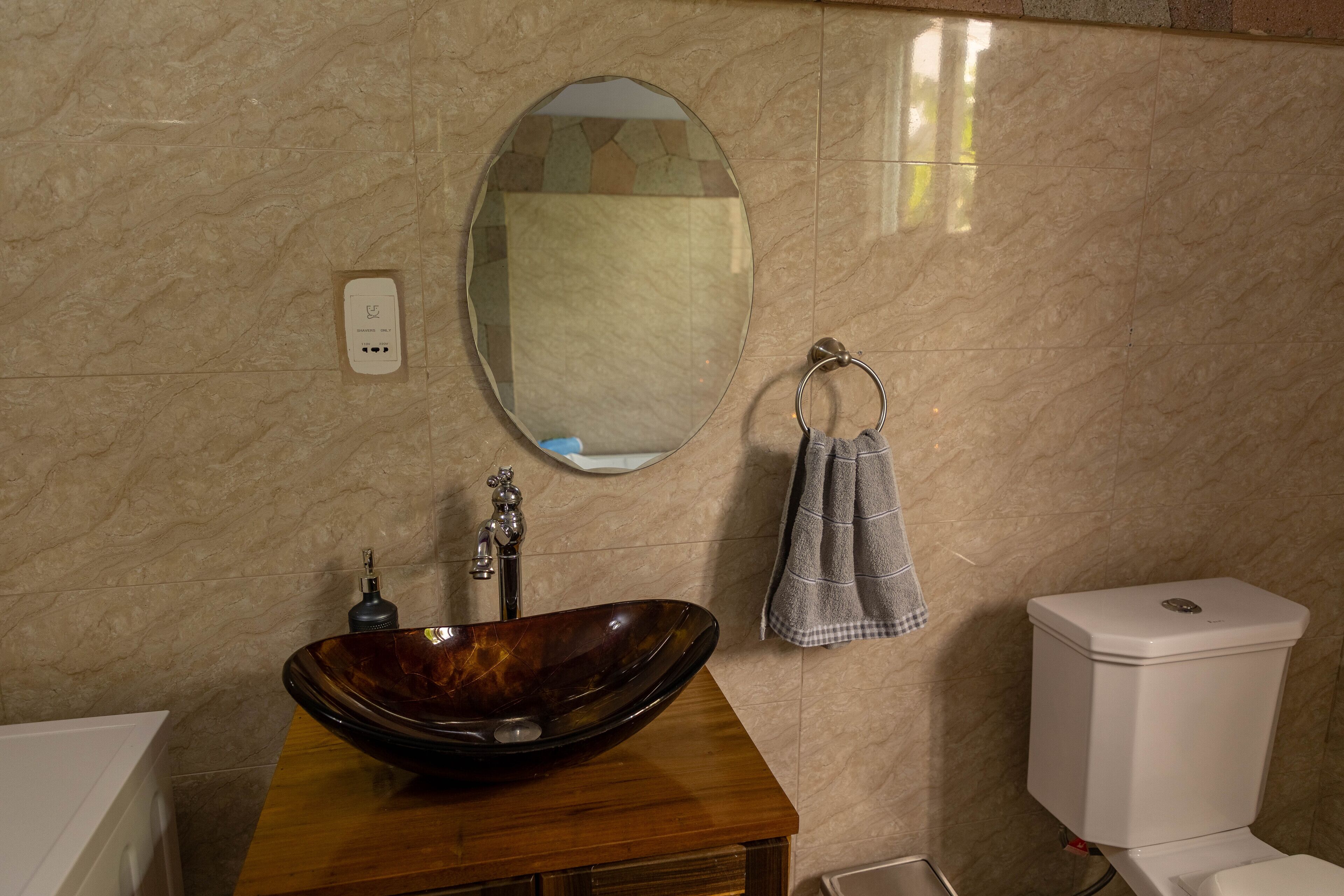 A bathroom with a wooden vanity, a vessel sink, a round mirror, a towel rack, and a toilet.