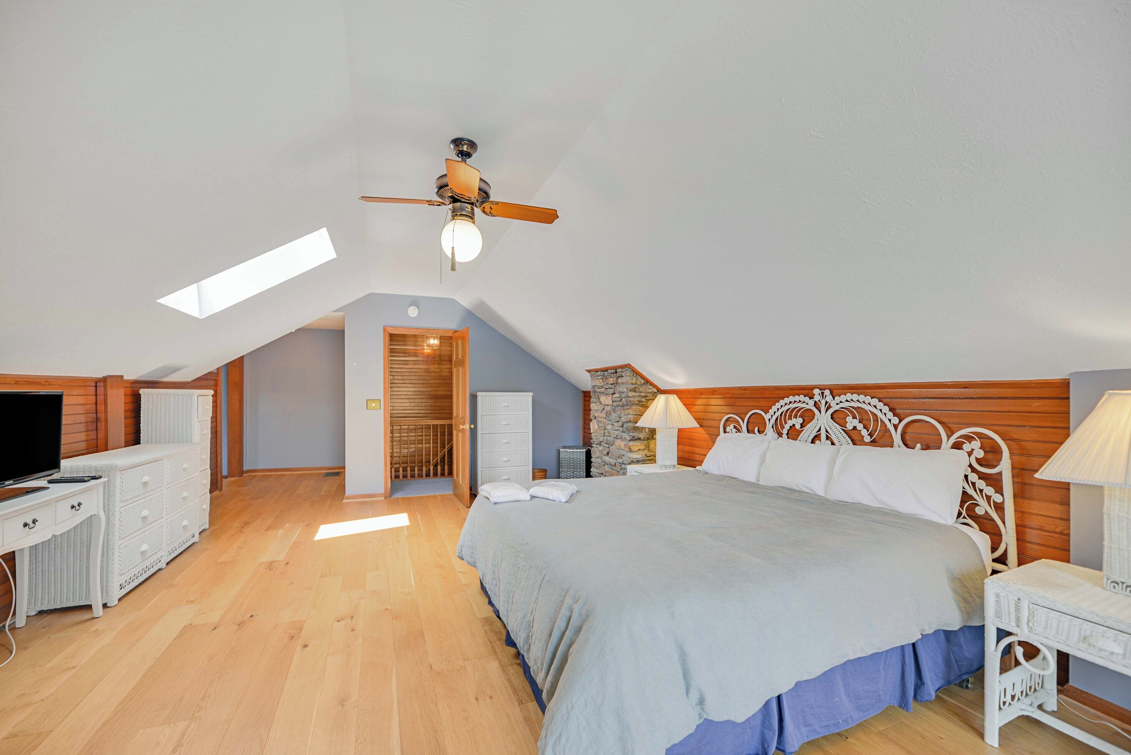 A bedroom with a large bed, a desk, a TV, and a ceiling fan.