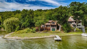 A lakeside property with a dock, a large weeping willow tree, and a house with multiple decks and a stone foundation.