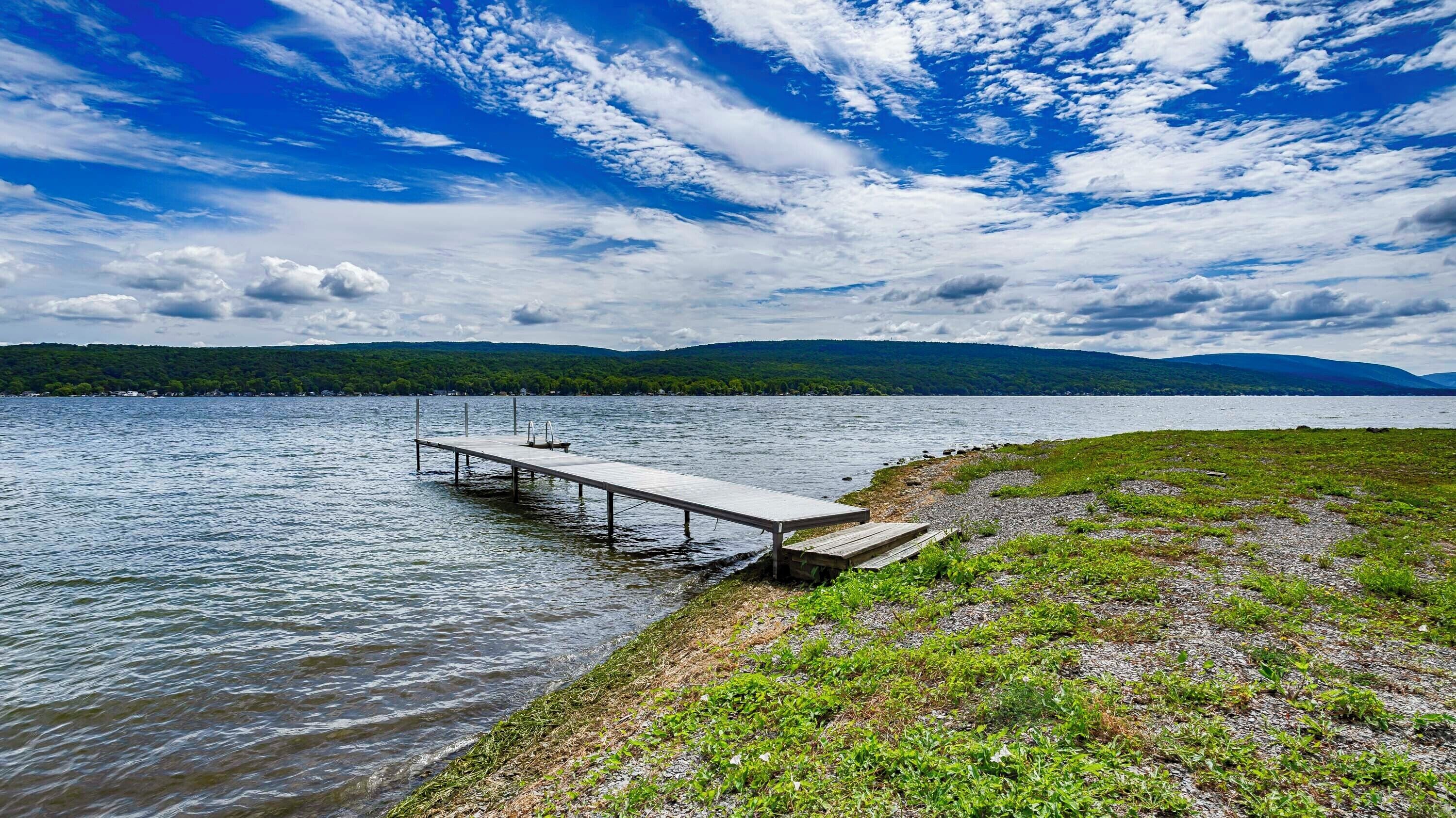 A lakeside dock extending into calm water, surrounded by grassy banks and distant mountains under a partly cloudy sky.