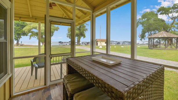 Un espacio de estar al aire libre techado con una mesa y bancos de madera, grandes ventanales con vistas a un área verde y un edificio distante.