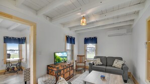 Amplia sala de estar con un sofá gris, una mesa de centro de madera y un televisor sobre un mueble de madera. Se observan dos ventanas con cortinas azules, una silla de madera y un sillón estampado.