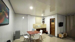 Un espacio habitable compacto con kitchenette, una zona de comedor con mesa y sillas, un televisor pequeño y una puerta que da a otra habitación.