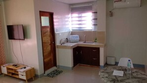 Una habitación pequeña y bien iluminada, con una puerta de madera, una pequeña kitchenette con microondas, un lavamanos y armarios. Hay un televisor montado en la pared, una mesa de madera pequeña y una mesa redonda de mármol con una botella de agua.