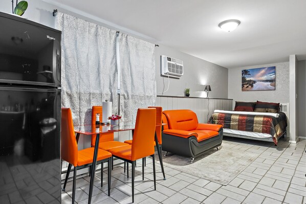 Een compact woongedeelte met een kitchenette, een eethoek met oranje stoelen, een bank en een bed.
